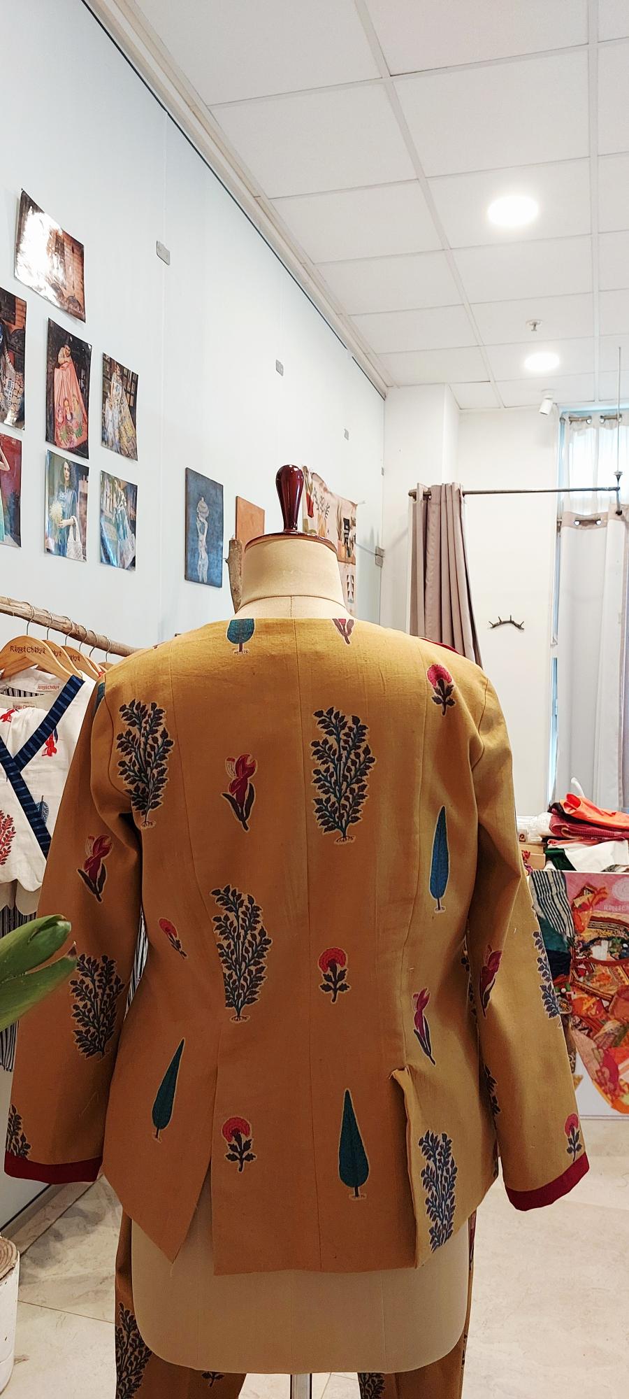 A mustard yellow embroidered jacket displayed on a dress form mannequin inside a clothing boutique. The jacket features colorful floral and leaf patterns in blue, red, and green, with red trim on the cuffs.