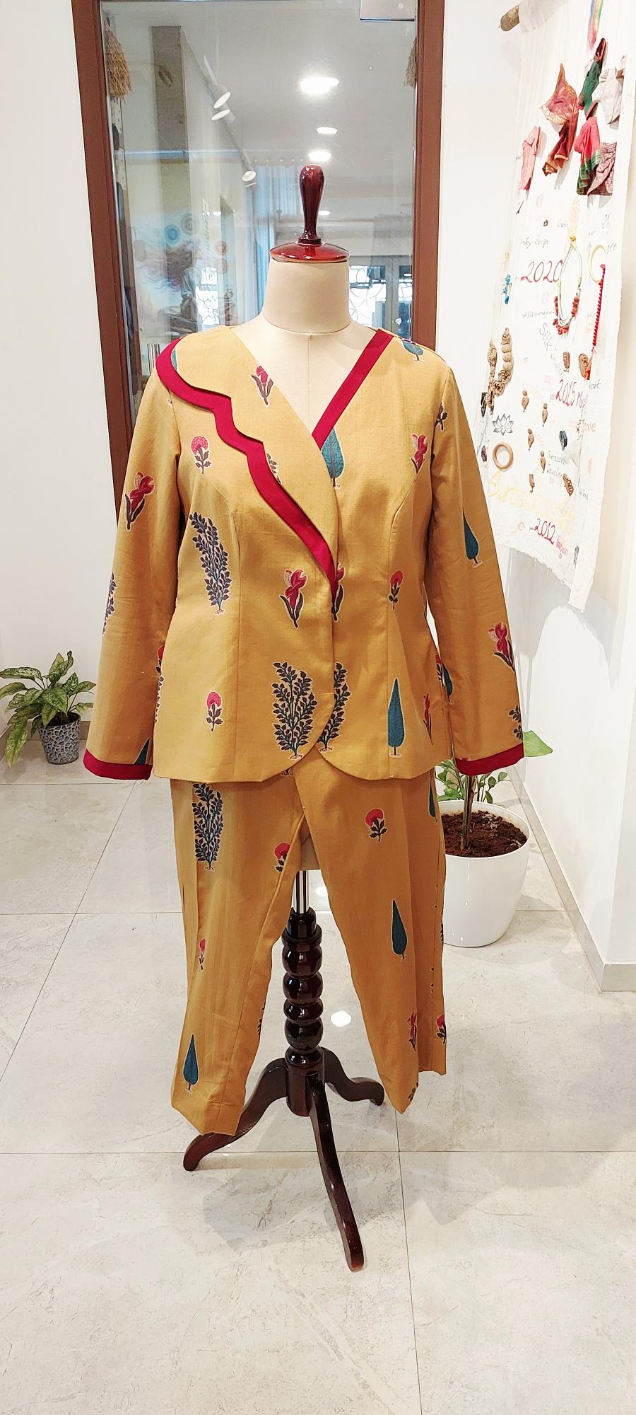 A mustard yellow two-piece traditional outfit with red and multicolor floral embroidery, displayed on a mannequin stand in an indoor boutique setting.